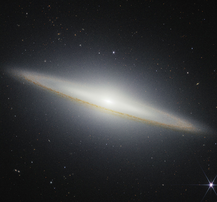 Image of a galaxy on the black background of space. The galaxy is a very oblong, brownish yellowish disk that extends from left to right at an angle (from about 10 o’clock to 5 o’clock). Mottled dark brown patches rim the edge of the disk and are particularly prominent where they cross directly in front of the galaxy. The galaxy’s center glows white and extends above and below the disk. There are different colored dots, distant galaxies, speckled among the black background of space surrounding the galaxy. At the bottom right, there is a particularly bright foreground star with Webb’s signature diffraction spikes.