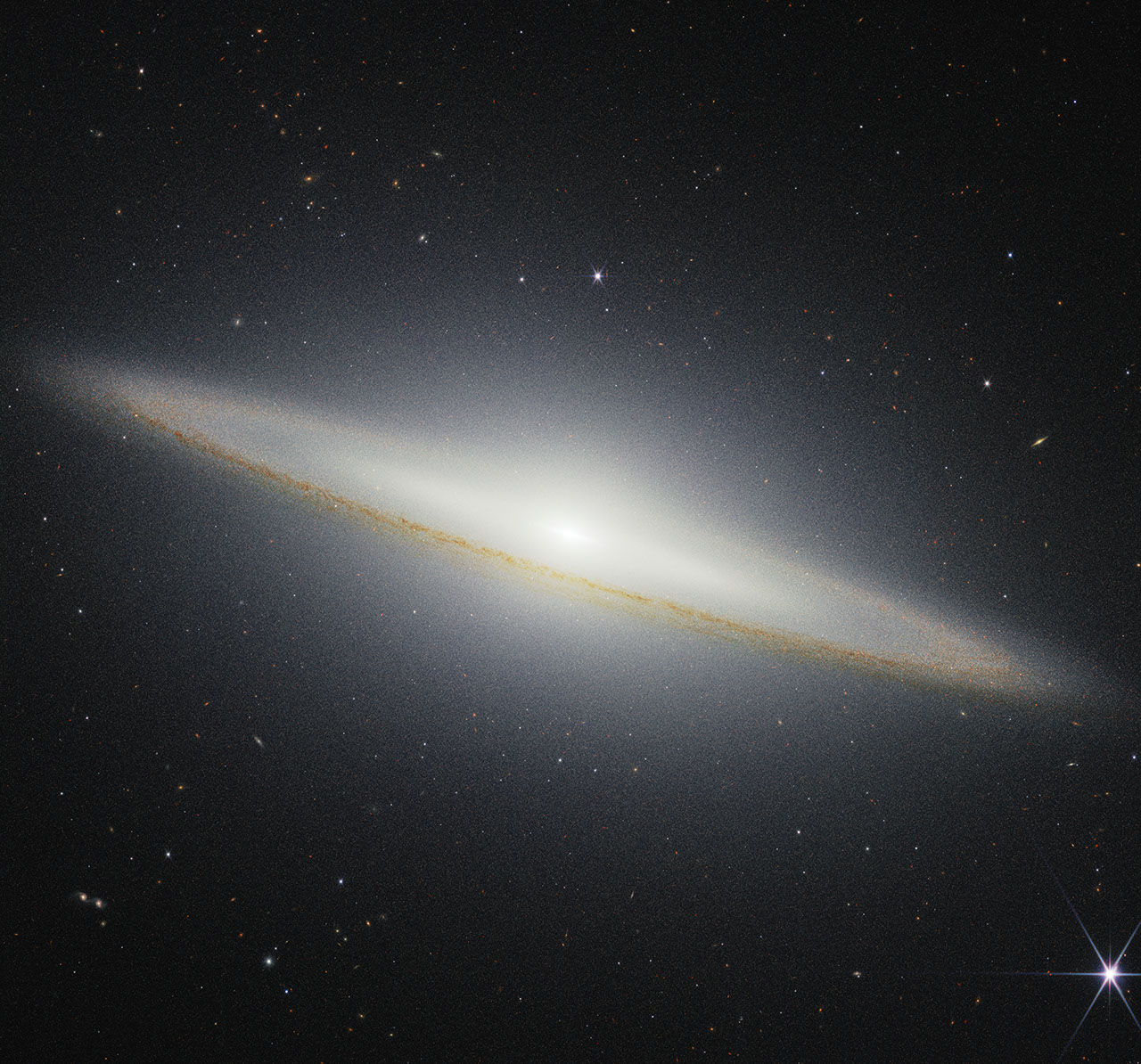 Image of a galaxy on the black background of space. The galaxy is a very oblong, brownish yellowish disk that extends from left to right at an angle (from about 10 o’clock to 5 o’clock). Mottled dark brown patches rim the edge of the disk and are particularly prominent where they cross directly in front of the galaxy. The galaxy’s center glows white and extends above and below the disk. There are different colored dots, distant galaxies, speckled among the black background of space surrounding the galaxy. At the bottom right, there is a particularly bright foreground star with Webb’s signature diffraction spikes.