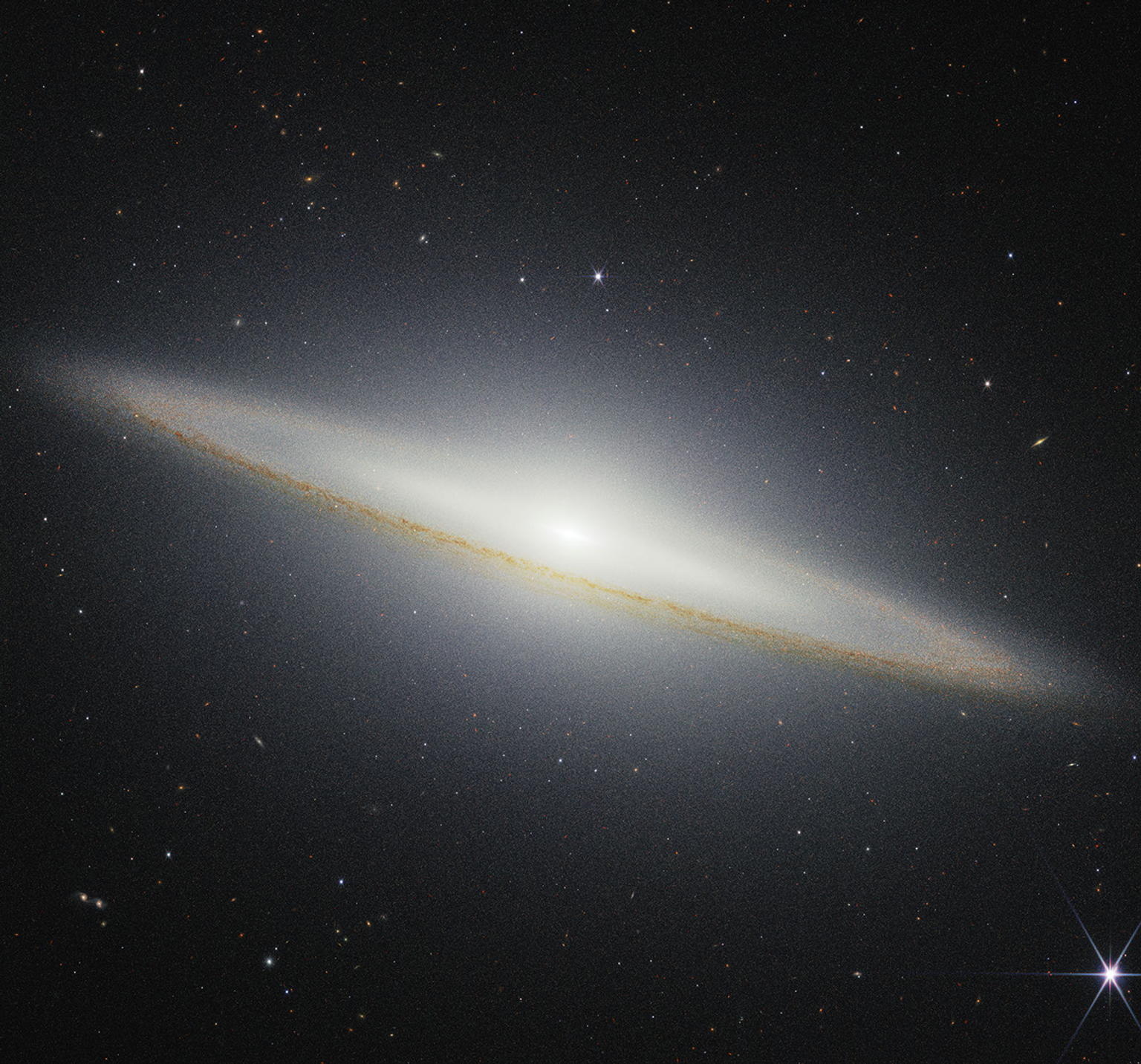 Image of a galaxy on the black background of space. The galaxy is a very oblong, brownish yellowish disk that extends from left to right at an angle (from about 10 o’clock to 5 o’clock). Mottled dark brown patches rim the edge of the disk and are particularly prominent where they cross directly in front of the galaxy. The galaxy’s center glows white and extends above and below the disk. There are different colored dots, distant galaxies, speckled among the black background of space surrounding the galaxy. At the bottom right, there is a particularly bright foreground star with Webb’s signature diffraction spikes.