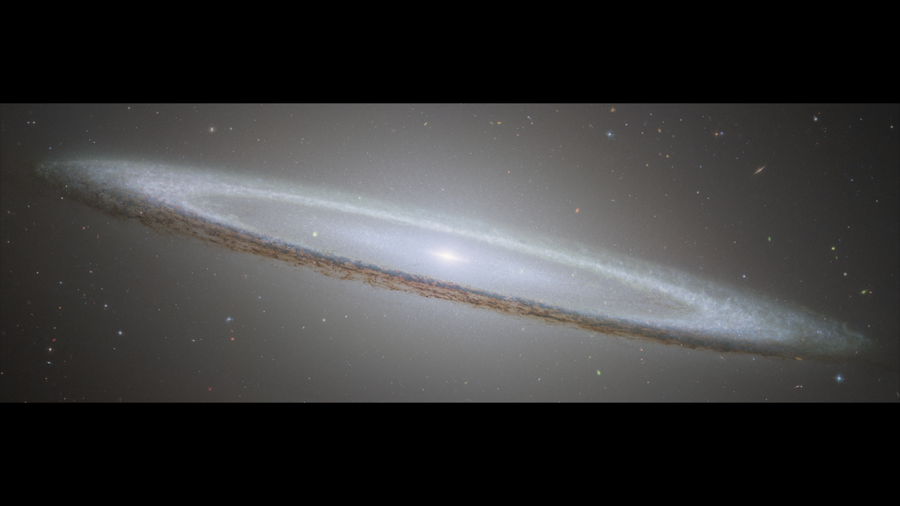 Hubble's image of the Sombrero galaxy. A very oblong, pale white disk of stars with a bright center extends from left to right on a black background. The disk is tilted so that the back side appears above the front side. Mottled dark patches rim the edge of the disk and are particularly prominent where they cross directly in front of the galaxy. It resembles a white dinner plate with a dark rim seen from the side, with the back tilted up and to the left slightly. A speckly white glow extends above and below the disk. Foreground stars also scattered across the scene.