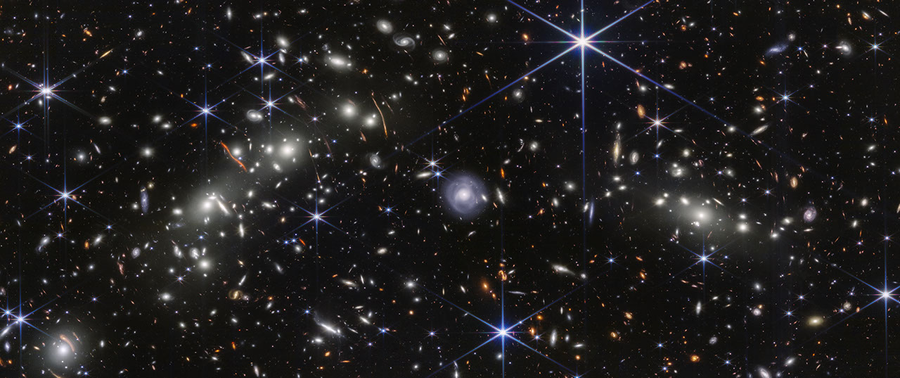 Webb observation of the Bullet Cluster shows many overlapping objects, including foreground stars, galaxies in galaxy clusters, and distorted background galaxies behind the galaxy clusters, set against the black background of space.