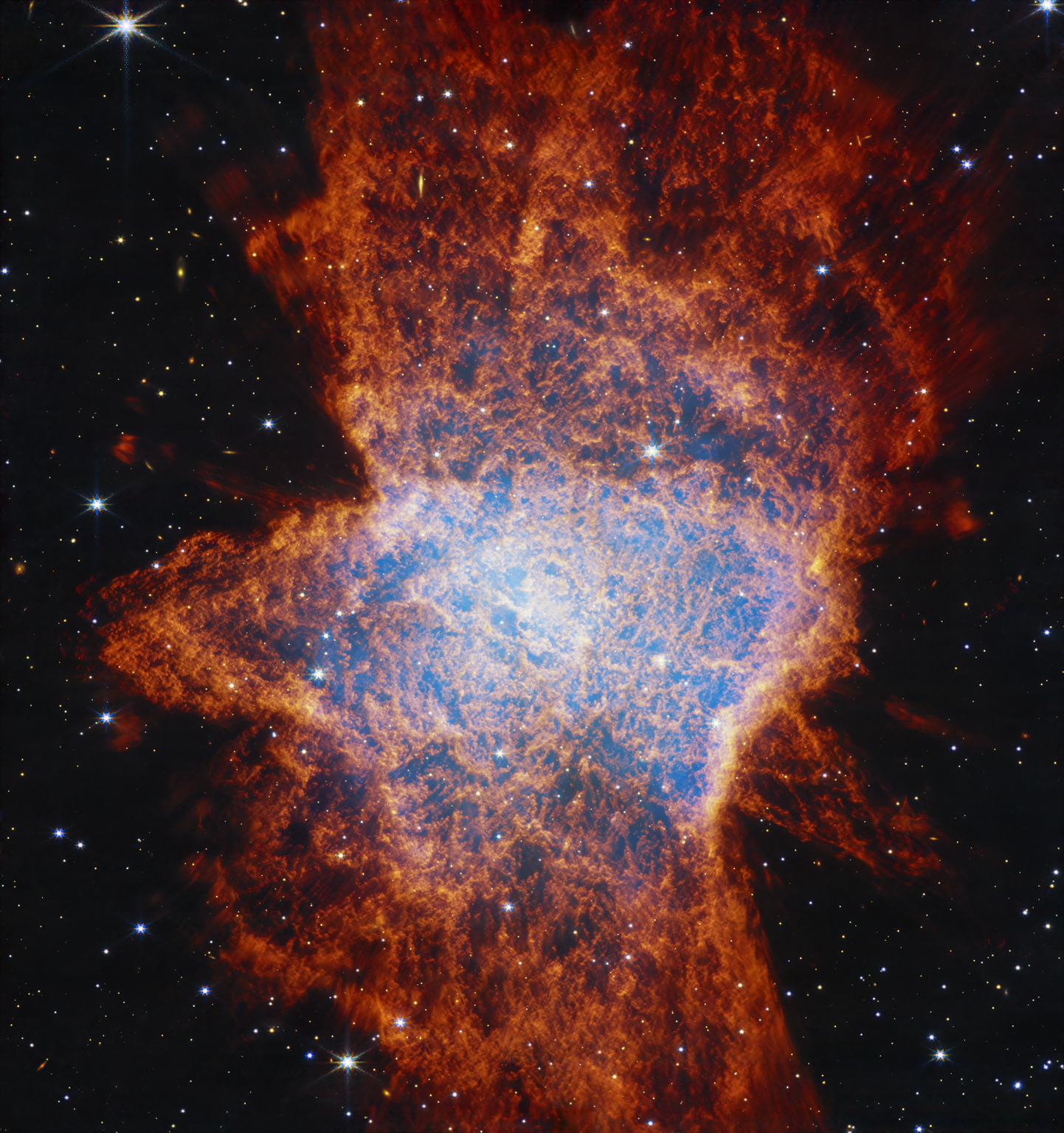 Colorful, mostly red glowing cloud with a distorted, asymmetrical shape that is illuminated from within by a bright central star. The asymmetrical shape resembles a large squished bug on the ground. In the center, a light blue glow appears over areas of dark pockets that look dark blue and are traced with orange material. It has a clumpy appearance. Shells of gas and dust appear as lobes stretching from roughly 11 to 5 o’clock, another from 1 to 7 o’clock, and possibly a third from 12 to 6 o’clock. The shells become a deeper red with distance from the center. These outflows push gas toward the equatorial plane, forming a disk that appears to span from 9 to 3 o’clock. The background of space is black and speckled with tiny bright stars and distant galaxies.
