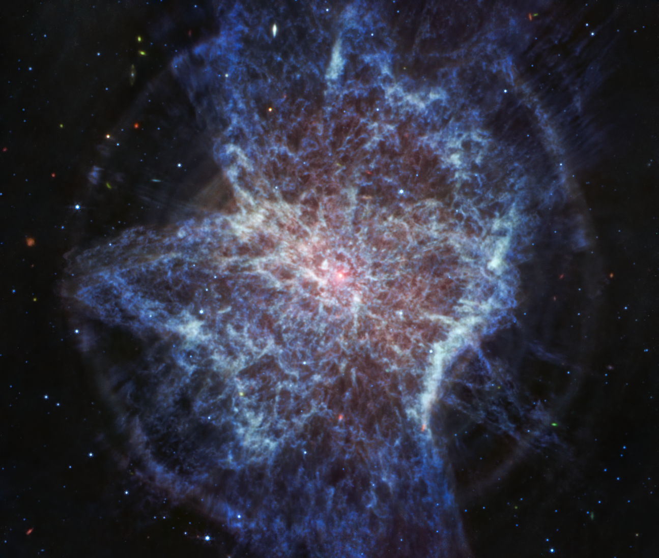 Colorful, mostly blue image of mid-infrared light from a glowing cloud with a distorted, asymmetrical shape. A star at the center of the image is a small point of pinkish-white light. The asymmetrical shape of the expanding cloud of gas and dust resembles paint splattered on the ground. The filaments of the expanding shells are wispy, and mostly white and blue. The shells appear as lobes stretching from roughly 11 to 5 o’clock, another from 1 to 7 o’clock, and possibly a third from 12 to 6 o’clock. These outflows push gas toward the equatorial plane, forming a disk that appears to span from 9 to 3 o’clock. A perfect circle of whitish blue dust traces the outer edges of the shells. The background of the image is black and speckled with tiny bright stars and distant galaxies.