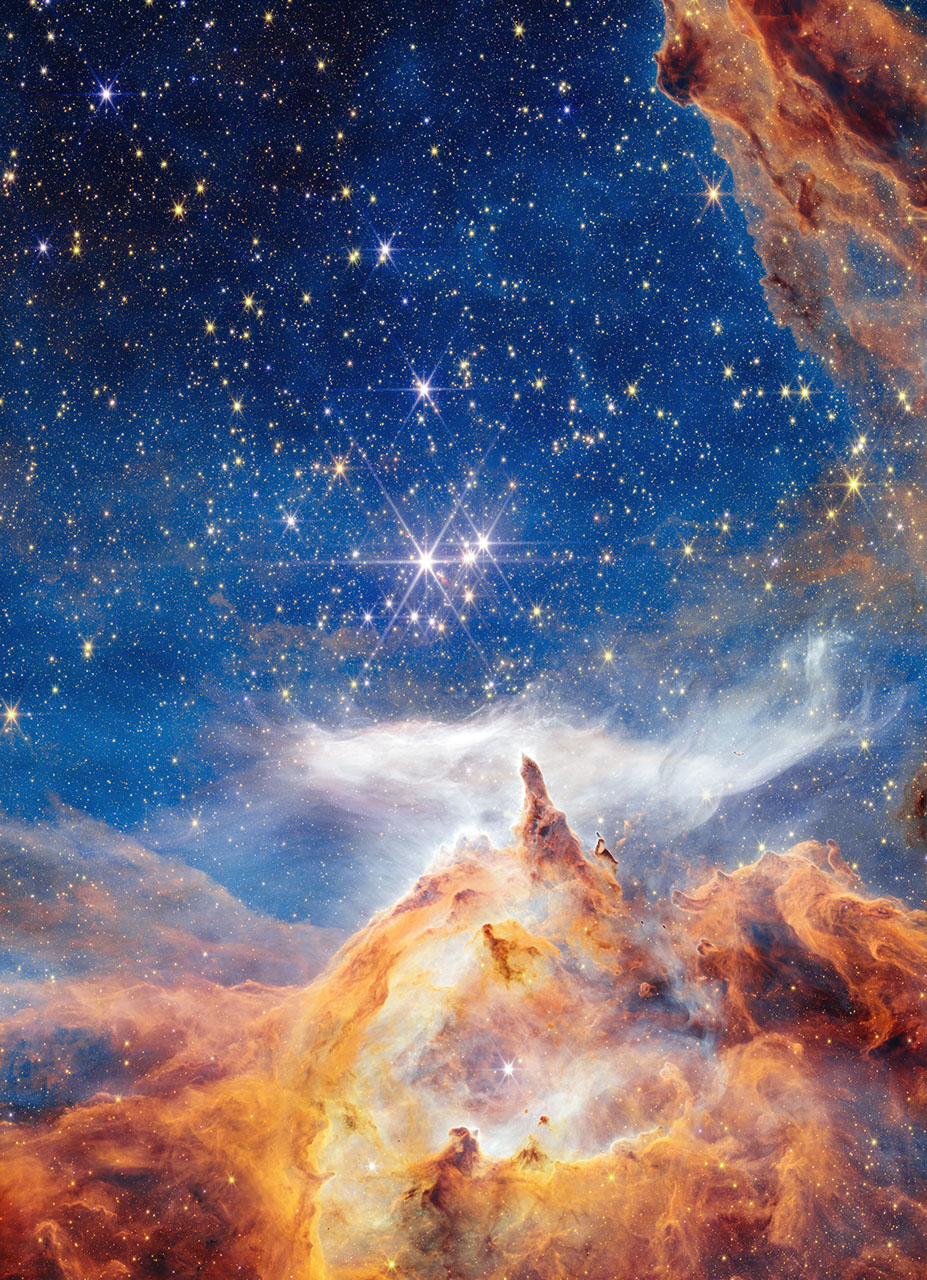 In what appears as a celestial dreamscape, a blue and black sky filled with brilliant stars covers about two thirds of the image. The stars are different sizes and shades of white, beige, yellow, and light orange. Across the bottom third of the scene is a craggy, mountain-like vista with spire-like peaks and deep, seemingly misty valleys. These so-called mountains appear in varying shades of orange, yellow, and brown. Above their soaring spires is a wispy, ethereal white cloud that stretched horizontally across the scene. Steam appears to rise from the mountaintops and join with this cloud. At the top, right corner of the image, a swath of orange and brown structure cuts diagonally across the sky.