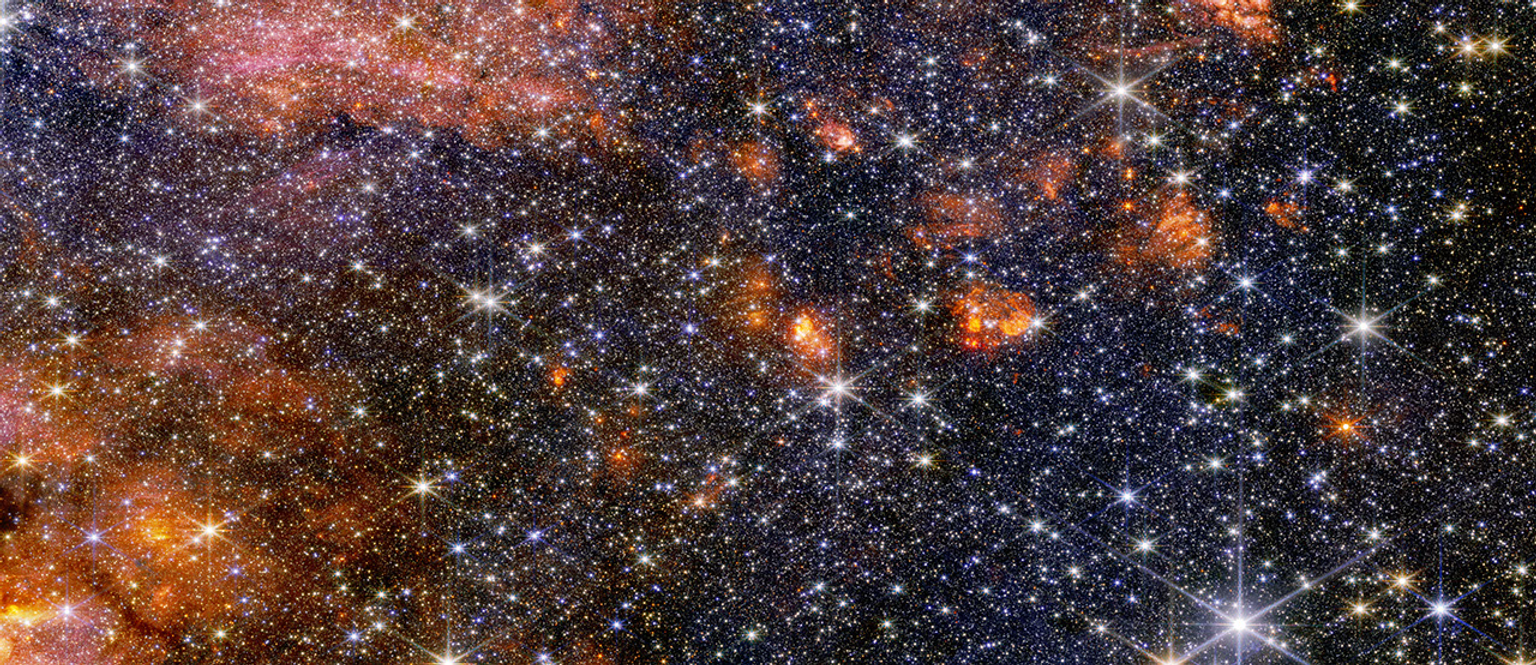 A wide view of a region of space filled with stars and clumps of orange clouds.
