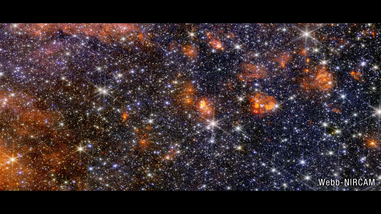 A wide view of a region of space filled with stars and clumps of orange clouds.