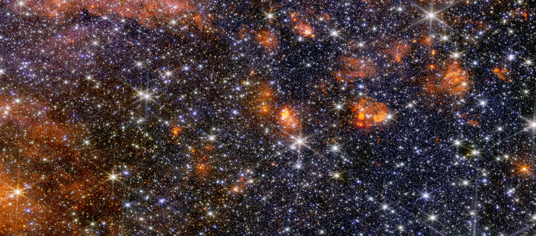 A wide view of a region of space filled with stars and clumps of orange clouds.