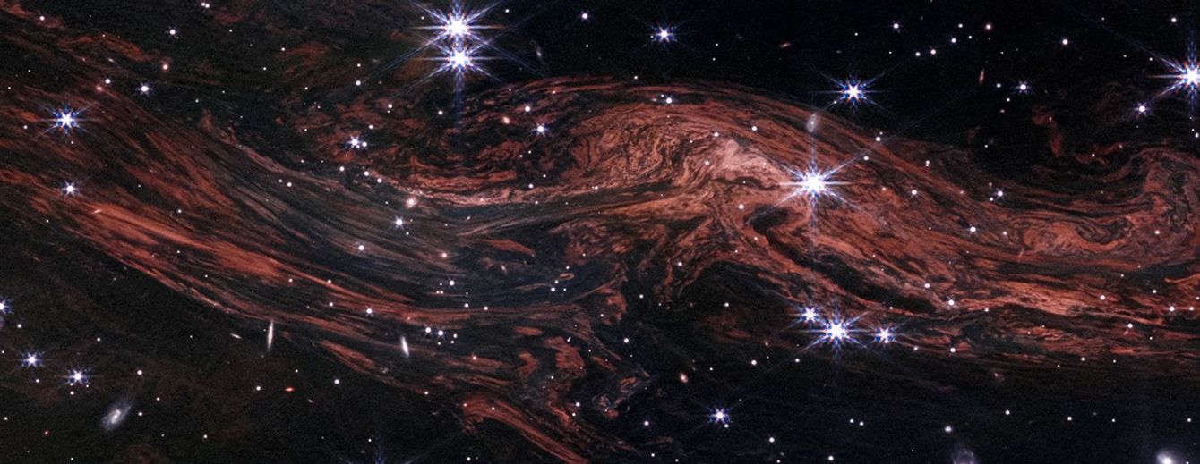 A few dozen white stars, some with Webb’s signature 8-point diffraction spikes, on the black background of space. Tightly packed, red-orange filaments extend horizontally from upper left to lower right, with a bulging upward curve on the right that is closer to the top. The filaments resemble wood grain.