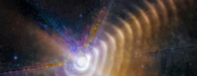 A portion of Webb’s 2023 observation of Wolf-Rayet 140. A bright white hexagon is toward the bottom left-center. This is the two stars. Blue diffraction spikes point diagonally toward 8, 11, and 1 o’clock. Surrounding the central light are a series of regularly spaced rings. A segment of each of the rings at around 2 o’clock appears brighter. These bright patches form a line that travels to the upper right. A few blue background dots are on the black background of space.