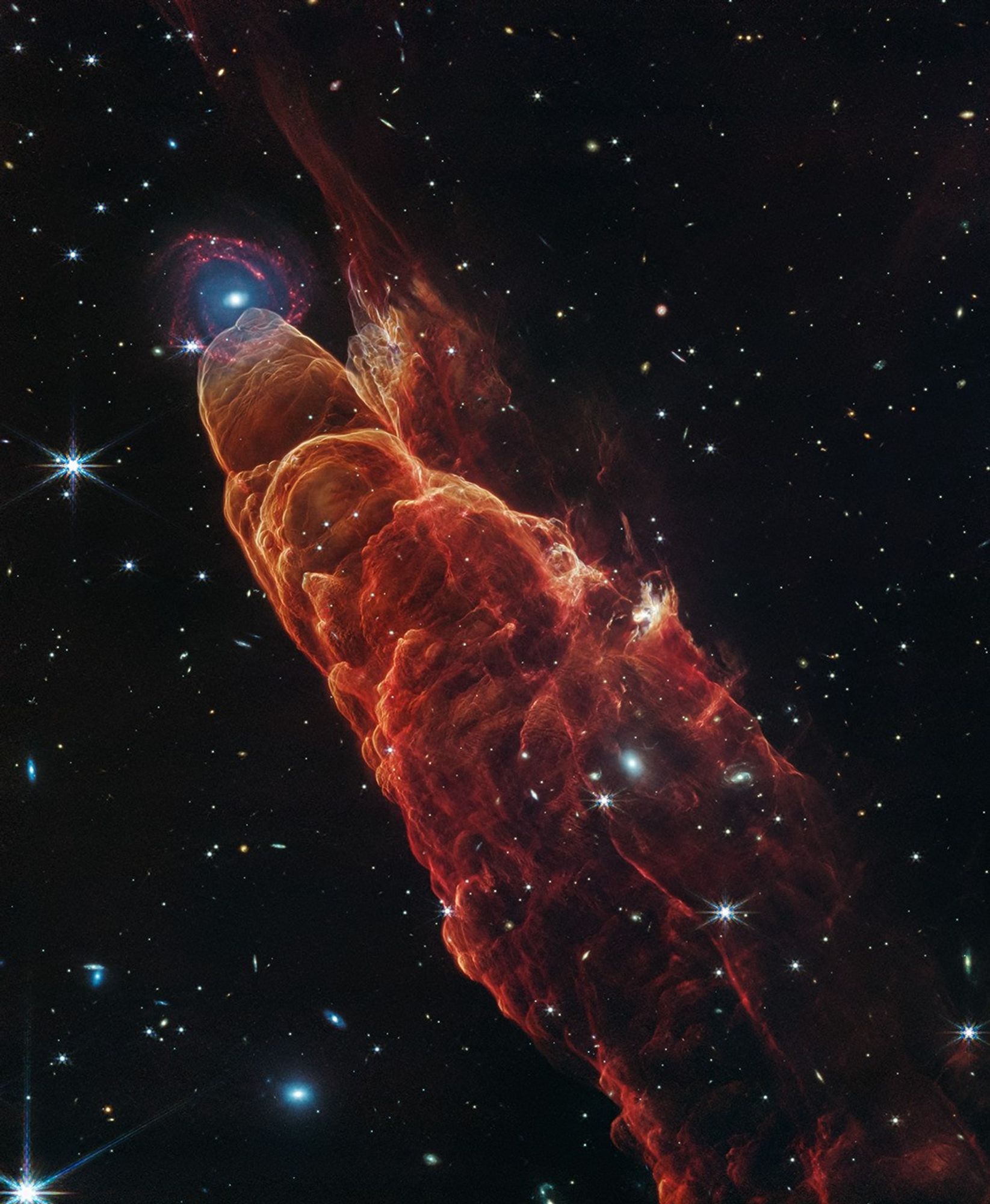 Angled from the upper left corner to the lower right corner of the image is a conical shaped orange-red cloud known at Herbig-Haro 49/50. This feature takes up about three-fourths of the length of this angle. The upper left end of this feature has a translucent, rounded end. At this same location there is background spiral shaped galaxy with a concentrated blue center that fades outwards to blend in with red spiral arms. The conical feature widens slightly from the rounded end at the upper right down to the lower right. Along the way there are additional rounded edges, like edges of a wave, intricate foamy-like details, as well as a clearer view of the black background of space. The black background of space is clearer, speckled with some white stars and smaller, more numerous, fainter white galaxies.