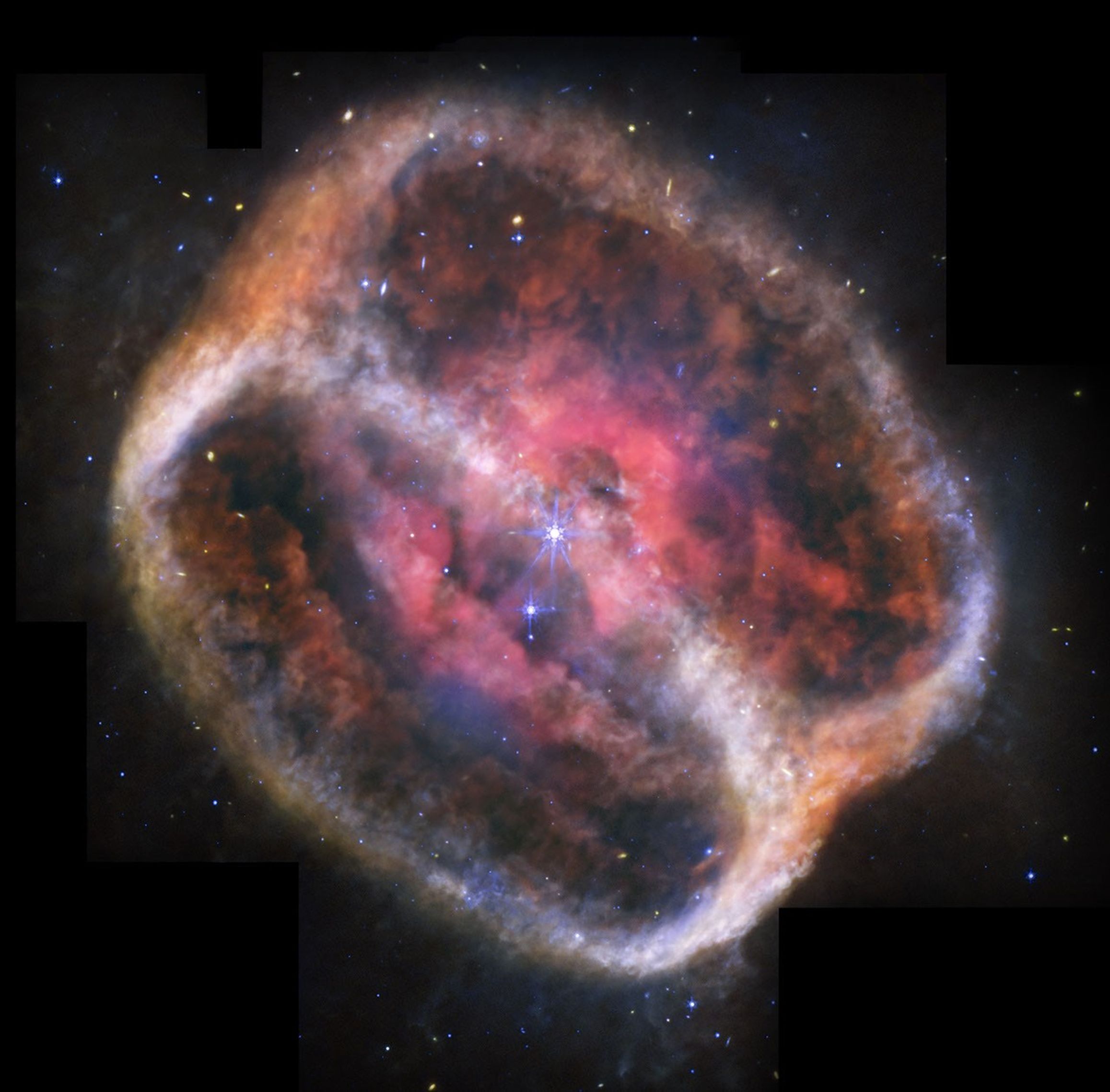 What looks like a single large, bright star (but is two) shines with bright purple diffraction spikes at the center of a large, diffuse cylinder of gas and dust that is tipped to the right. At the center is a bright pink clumpy cloud that takes up about 25% of the view. The pink region has some holes and diffuse areas. Beyond that are two large rings seen at a roughly 60-degree angle that appear joined at top left and bottom right. The edges are denser, and form shallow V-shapes that go inward. The rings appear orange at top left and bottom right, and are blue at bottom and center right. There is diffuse orange material around the body. The black background of space is speckled with tiny stars and galaxies mostly in blues and yellows. A bigger blue star with spikes is just below and to the left of the central stars, but it is slightly smaller. Areas Webb did not observe are along the top edges, a thin vertical near the nebula at top left, and at the bottom left and right corners.