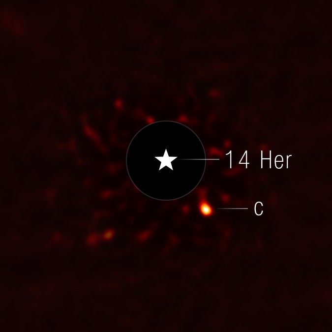 This image shows the exoplanet 14 Herculis c. The view is mostly black, with very faint red splotches in the central region of the image. At the center of the image, there is a black circle, and in the center of that, there is a star symbol representing a real star. This black circle blocks the light from the host star. To the lower right of the circle is a fuzzy bright orange circle, which is the exoplanet.