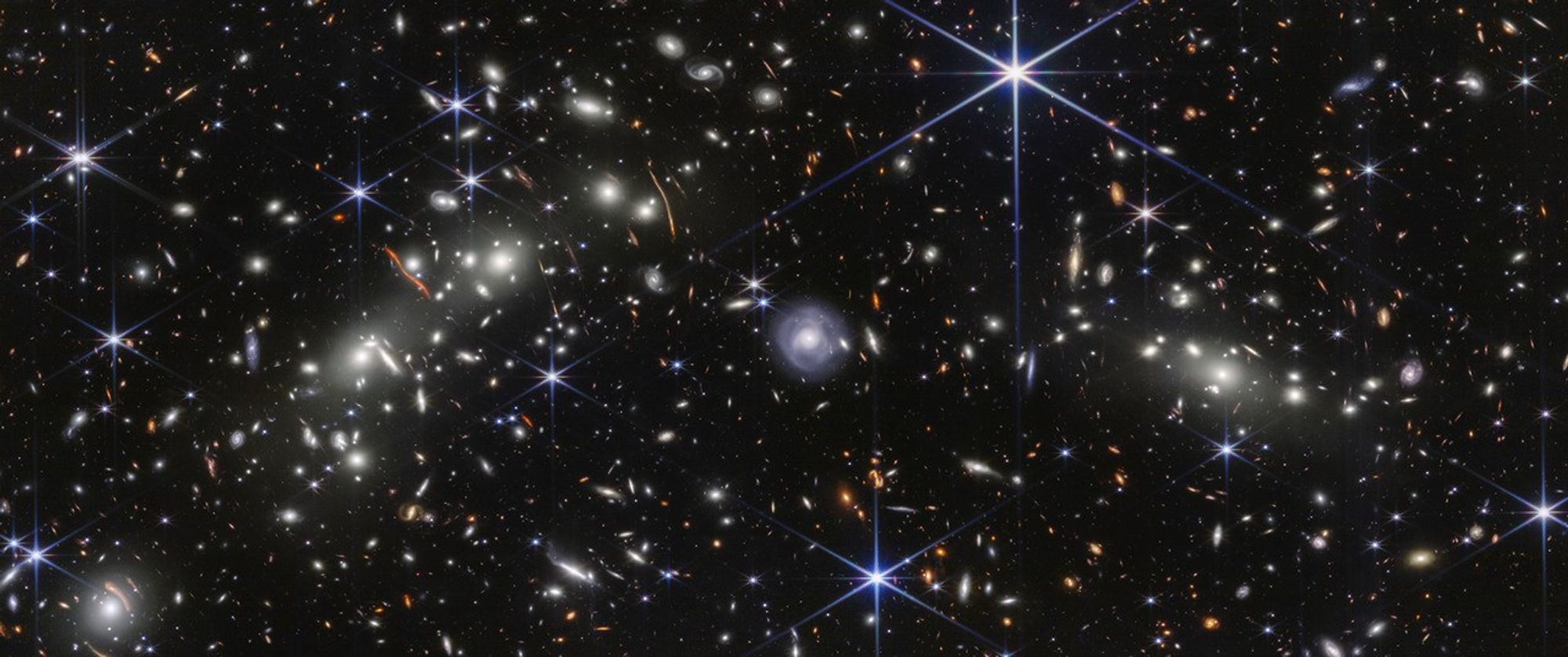 Webb observation of the Bullet Cluster shows many overlapping objects, including foreground stars, galaxies in galaxy clusters, and distorted background galaxies behind the galaxy clusters. The objects are all at various distances set against the black background of space. Most galaxies appear as fuzzy ovals in white, orange, or red, but a few have distinctive spiral arms. A slightly larger, very bright light blue spiral galaxy is at center. To its left are several bright white blob-like galaxies that are members of one galaxy cluster. On the opposite side of blue spiral galaxy is another, slightly smaller set of bright white galaxies, part of the second galaxy cluster. There are many thin, long, orange arcs that follow invisible concentric circles. These orange arcs, easiest to spot on the left, are background galaxies that have been stretched and distorted by the foreground galaxy clusters.