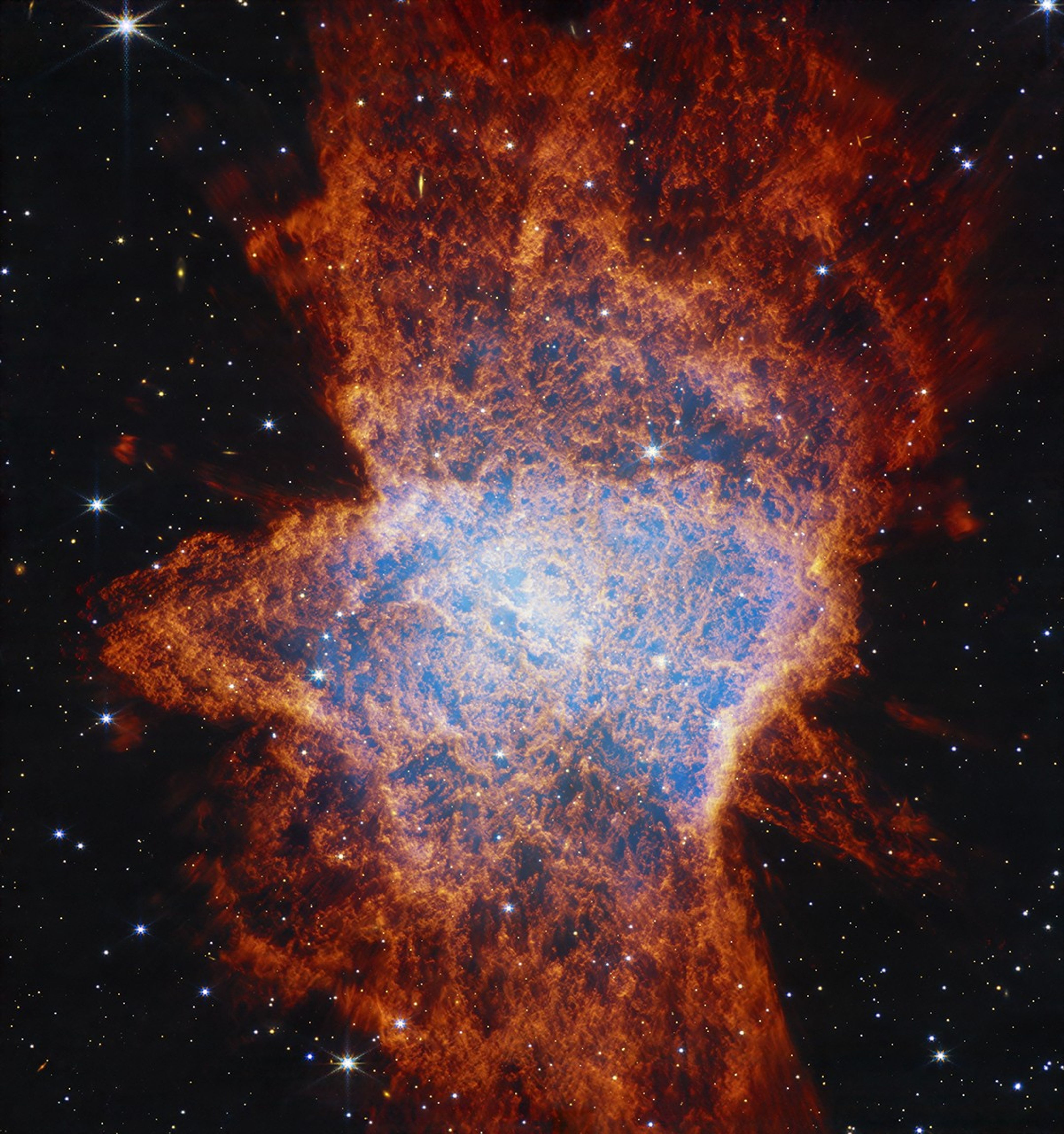 Colorful, mostly red glowing cloud with a distorted, asymmetrical shape that is illuminated from within by a bright central star. The asymmetrical shape resembles a large squished bug on the ground. In the center, a light blue glow appears over areas of dark pockets that look dark blue and are traced with orange material. It has a clumpy appearance. Shells of gas and dust appear as lobes stretching from roughly 11 to 5 o’clock, another from 1 to 7 o’clock, and possibly a third from 12 to 6 o’clock. The shells become a deeper red with distance from the center. These outflows push gas toward the equatorial plane, forming a disk that appears to span from 9 to 3 o’clock. The background of space is black and speckled with tiny bright stars and distant galaxies.