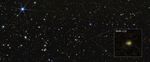 A wide field of view showing deep space, dotted with many small galaxies and a few foreground stars that display six diffraction spikes. One galaxy is highlighted with a magnified image in a graphic pull-out box in the lower right corner. The galaxy is labeled MoM-z14 and appears as a blurry yellow blob with a small red area at its top.