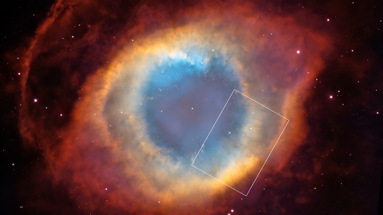 Almost resembling an eye, a circular region of glowing blue gas is ringed in yellow gas, surrounded by a football-shaped red region. The nebula shines against black space dotted with stars. There is a rectangular box outlined in white at about 4 o'clock.