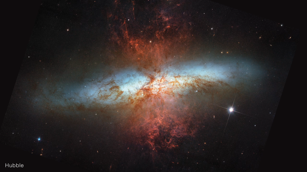 Edge-on spiral starburst galaxy Messier 82 as imaged by NASA's Hubble Space Telescope. Hourglass-shaped red-orange plumes of material are shooting outward from above and below a bright blue-white, disk-shaped center. Messier 82 is set against the black background of space, which has many distant galaxies that appear as small white and orange ovals and points of light. Toward the right of Messier 82 is a white star with four-pointed diffraction spikes that are characteristic of Hubble. Text in the bottom left corner reads u201cHubble.u201d