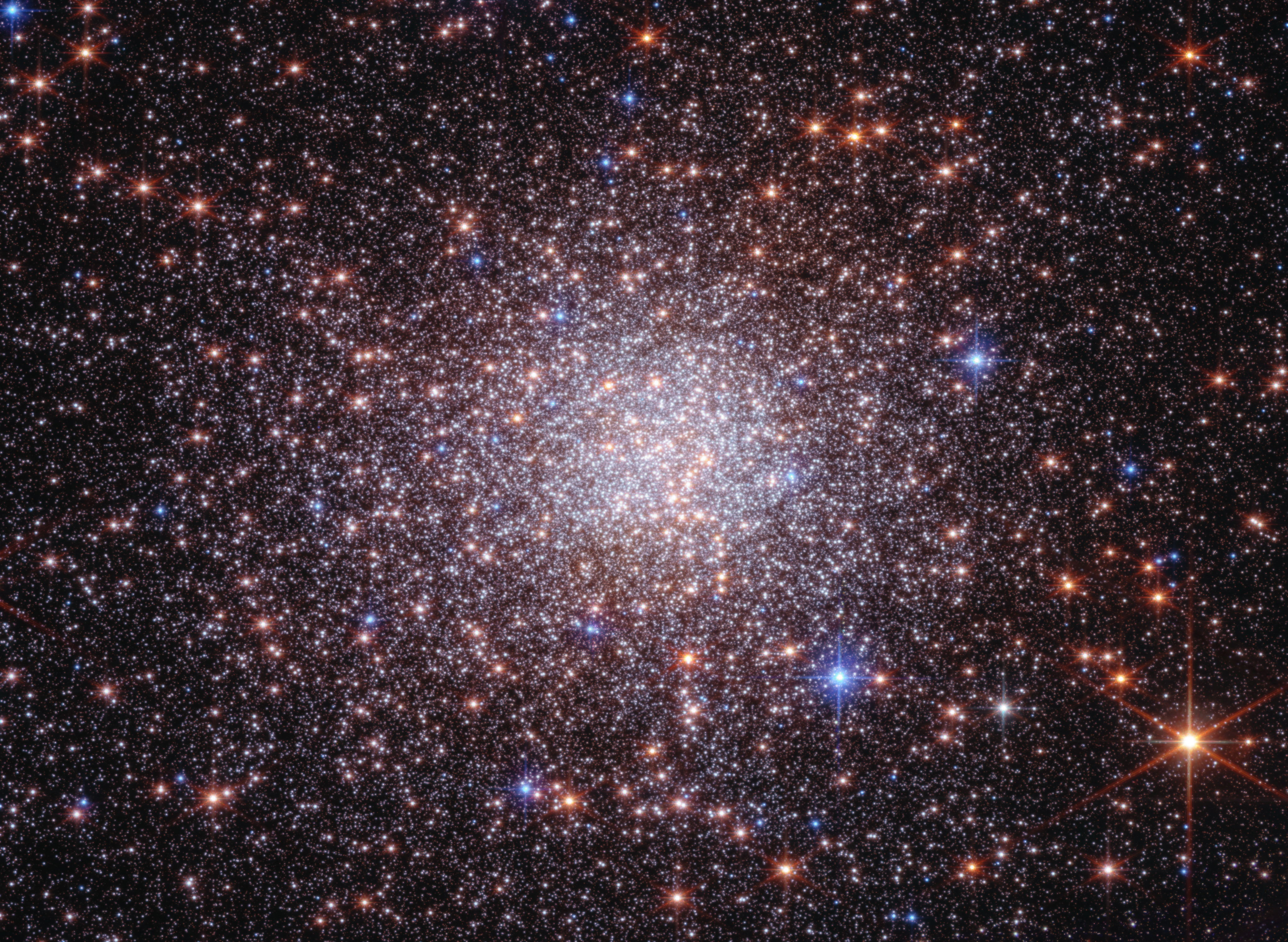 A dramatically crowded starfield that looks like a just-shaken snow globe. The black background of space, which is clearer at the edges, is covered by thousands of tiny white, orange, and blue points of light, which are stars. The stars are most concentrated in the center, forming a roughly circular orb, and sparser at the edges of the image. Several larger orange stars, particularly those largest near the edges of the frame, have prominent diffraction spikes.