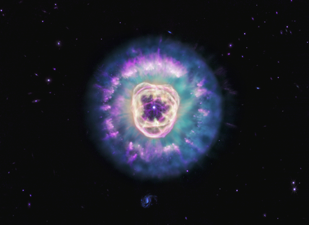 Planetary nebula NGC 2392, also called the Lion Nebula, against the black background of space. The nebula is in the center, circular in shape, and looks like a male lion’s head. In the very center is a small, bluish-white circle with eight-point diffraction spikes, a white dwarf star. Surrounding the star are light purple-red cavernous bubbles and shell-like rings. The bubbles and shells collectively form an oval with two small, wide arcs near the top, reminiscent of a lion’s face and its ears. What appears to be extended outward from the lion’s face is a thick ring of cyan material. The width of the ring is consistent throughout and resembles a mane. The area closest to the lion’s face looks cloudy, while the edges of the mane look like there are purple clumps of dust with comet-like tails and strands. In the background are distant galaxies and stars. Some galaxies appear as small purple points of light and others with visible blue spiral structures. A couple of the stars have diffraction spikes.