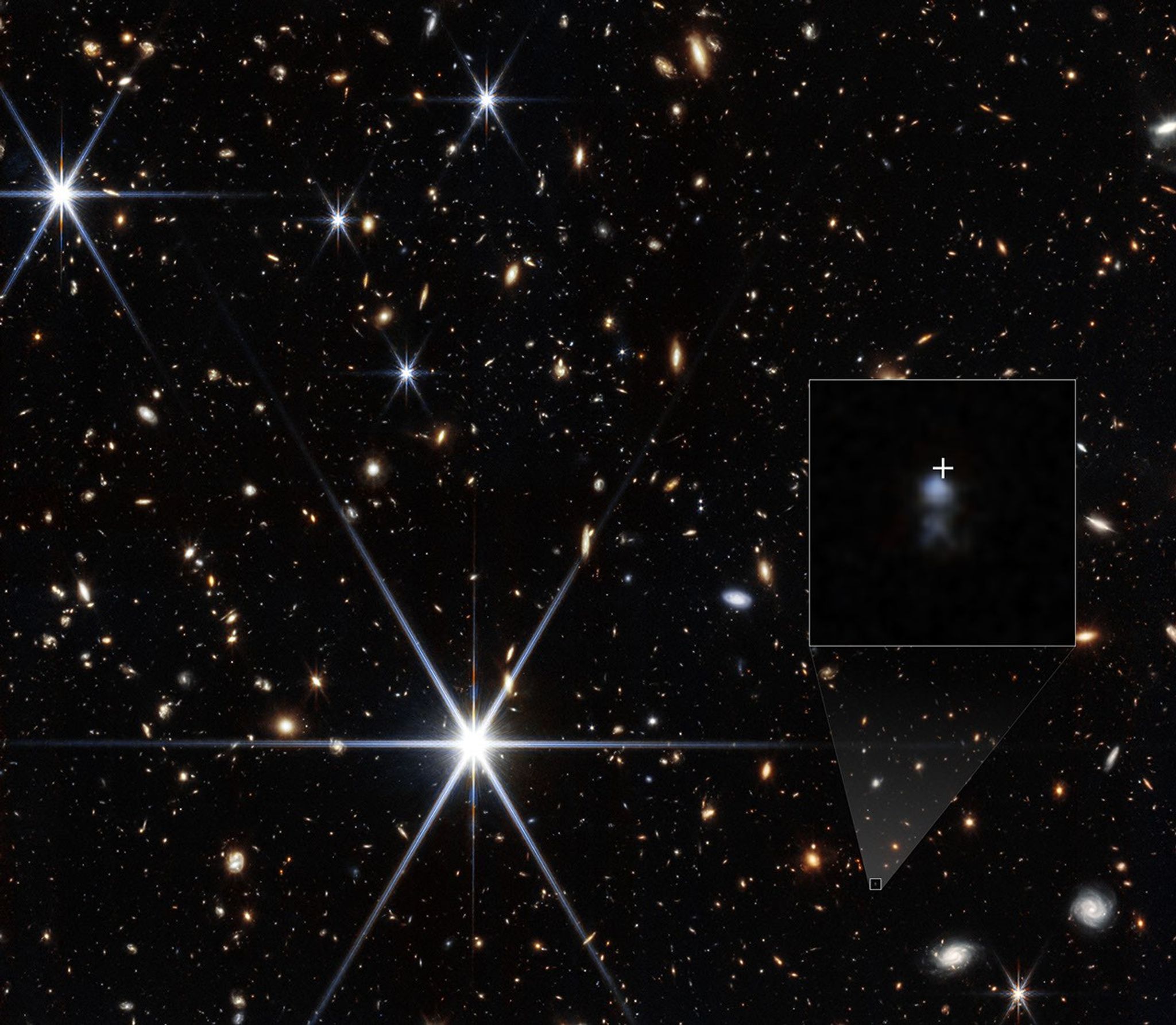A field full of small galaxies of all shapes against the blackness of space. A few bright white stars show 8 prominent diffraction spikes. A small galaxy at lower right is highlighted with a white box. A shaded triangle extends upward to the corners of a larger box showing a zoom in of the region. Two fuzzy, light blue blobs are at center, with a larger and brighter one at top. Just above the top blob is a white crosshair showing the location where a fast radio burst was detected.