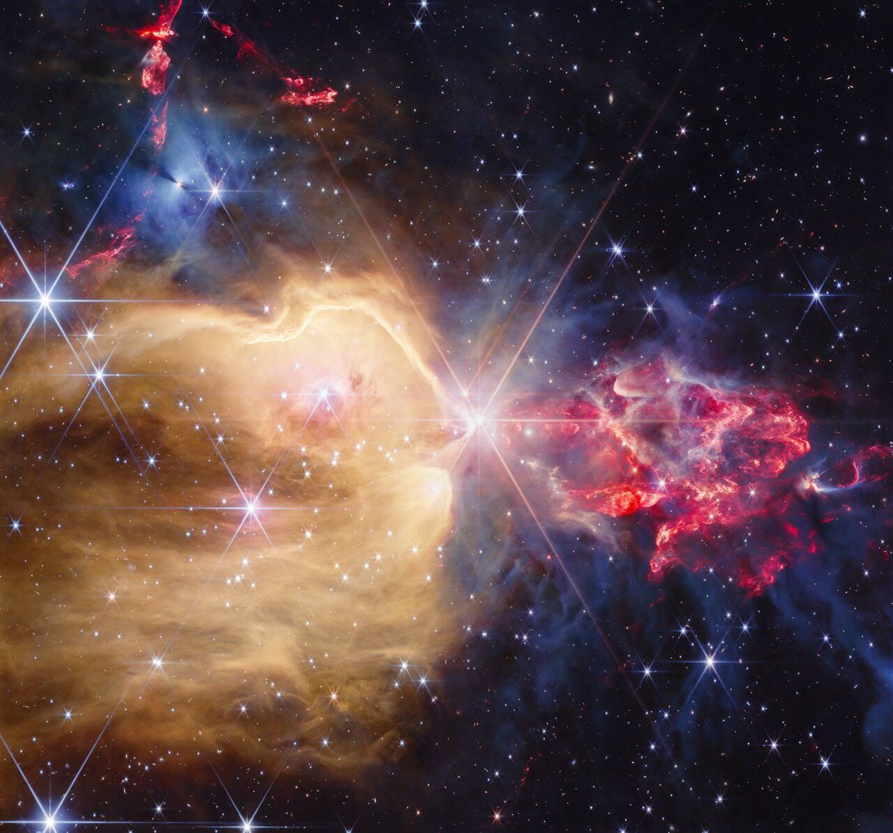 A young star cluster filled with many stars that display Webb’s unique eight-pronged diffraction pattern. At its center is a yellow star that sports the largest diffraction pattern. To its left, there is a region filled with yellow dust and gas that extends from the star to the left and the bottom, covering about two-thirds of the frame. The yellow region has several embedded blue stars of different sizes. The top third, which lies outside the yellow region, has a few bright protostars, within dense gray gas. These stars illuminate the gas, making it appear blue. A few red outflows from other protostars are visible above this grayish-blue region. To the right of the central star, there is a clumpy, flame-like plume of red dust and gas with protostars that emit a soft white light within. The plume is surrounded by more dense gray gas and dust. It is about one third the size of the yellow region. There are several background galaxies strewn throughout.
