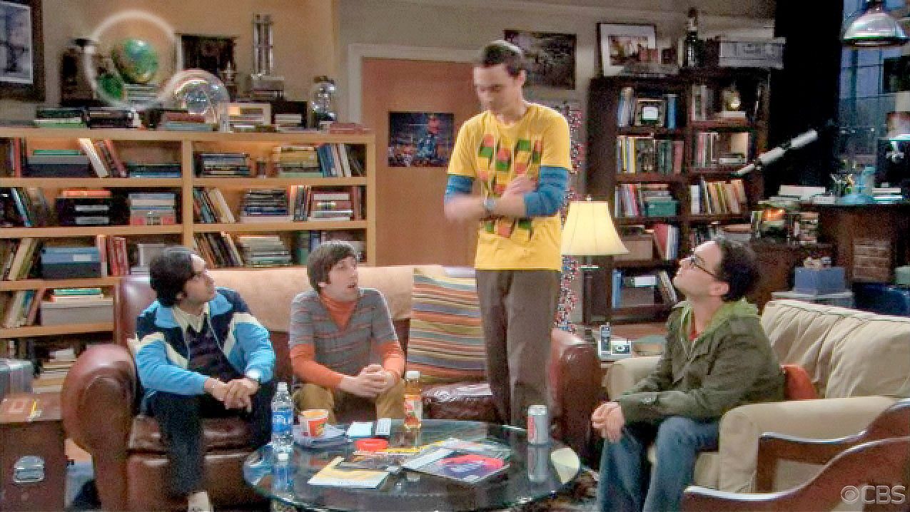 Scene from the TV show The Big Bang Theory, with four main characters in their apartment living room. One character stands while the others sit around a coffee table with drinks and papers. A colorful Hawking’s Beach Ball, showing the cosmic microwave background map, is visible on the bookshelf behind them.