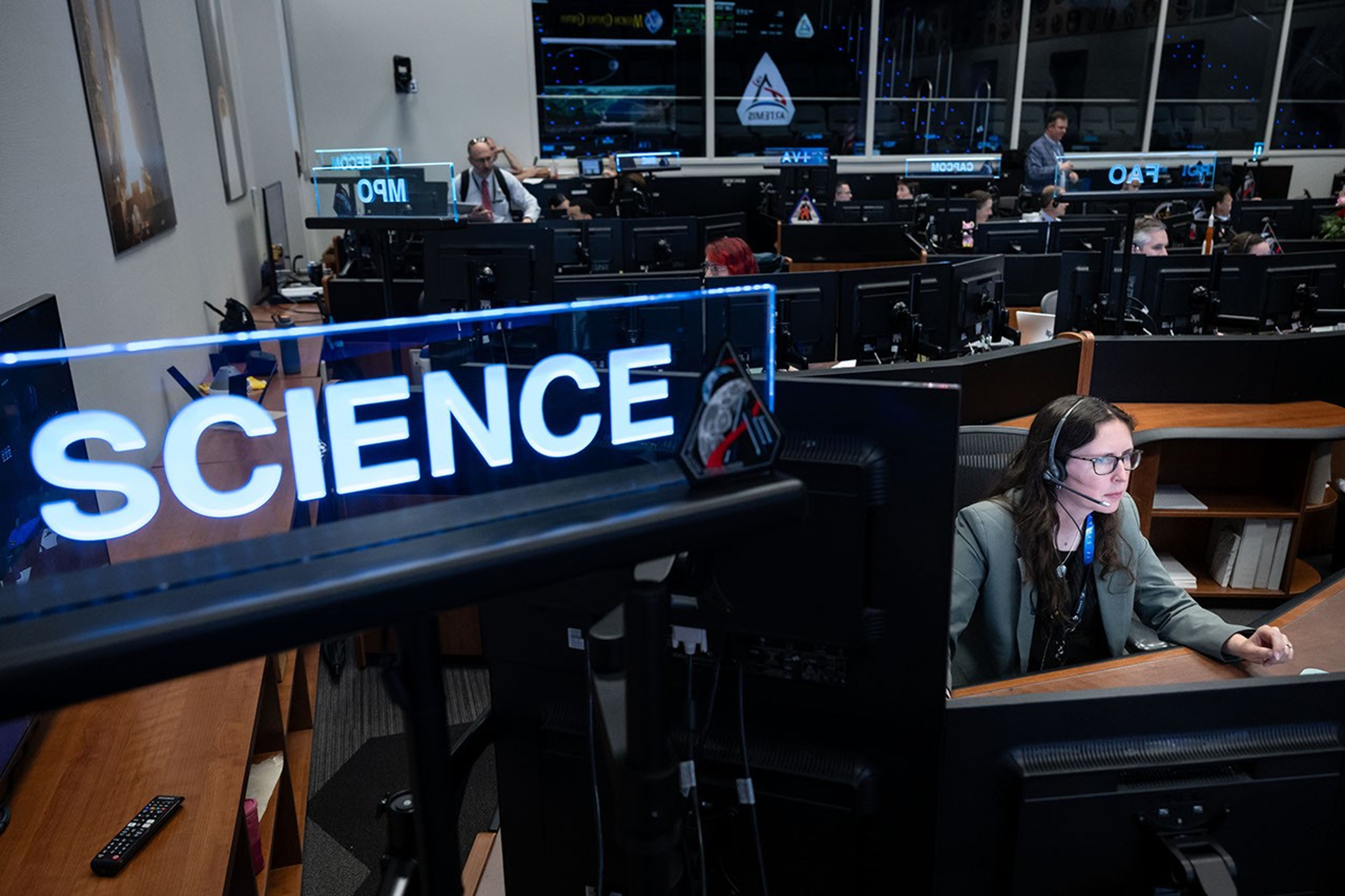 A picture of a woman sitting at a desk in room filled with large, modern, wooden desks that have multiple monitors across all of them. She is wearing business attire and is focused on a computer screen. She has long, dark, hair and is wearing glasses and a headseat with a microphone. On the left side of the image is a glass sign above the desk that reads, "SCIENCE."