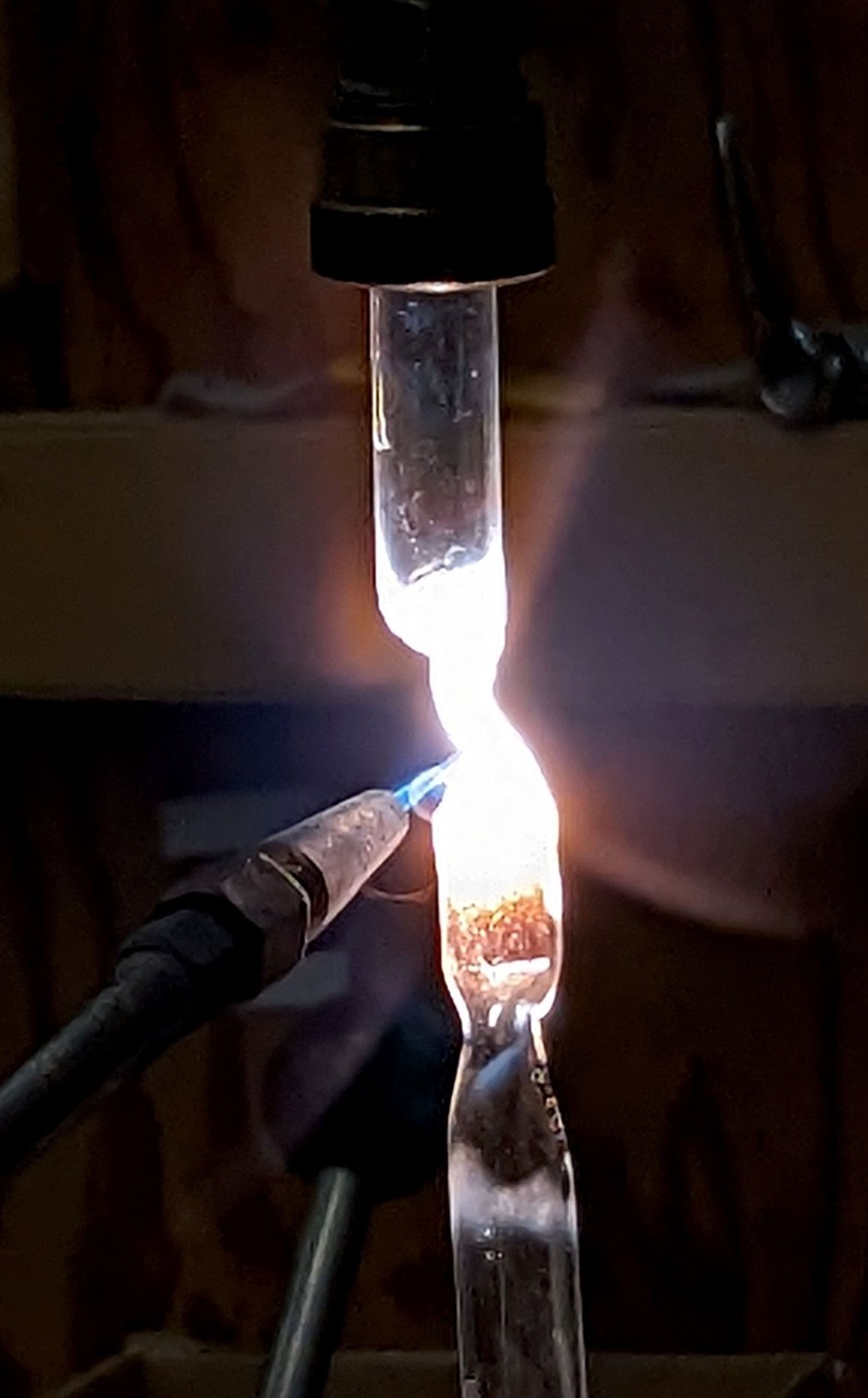 An image of a blue flame from a torch melting a glass tube shut. The image is mostly dark, with the glass tube coming right down the center. The middle of the tube is filled with a bright white glow from the glass being melted.
