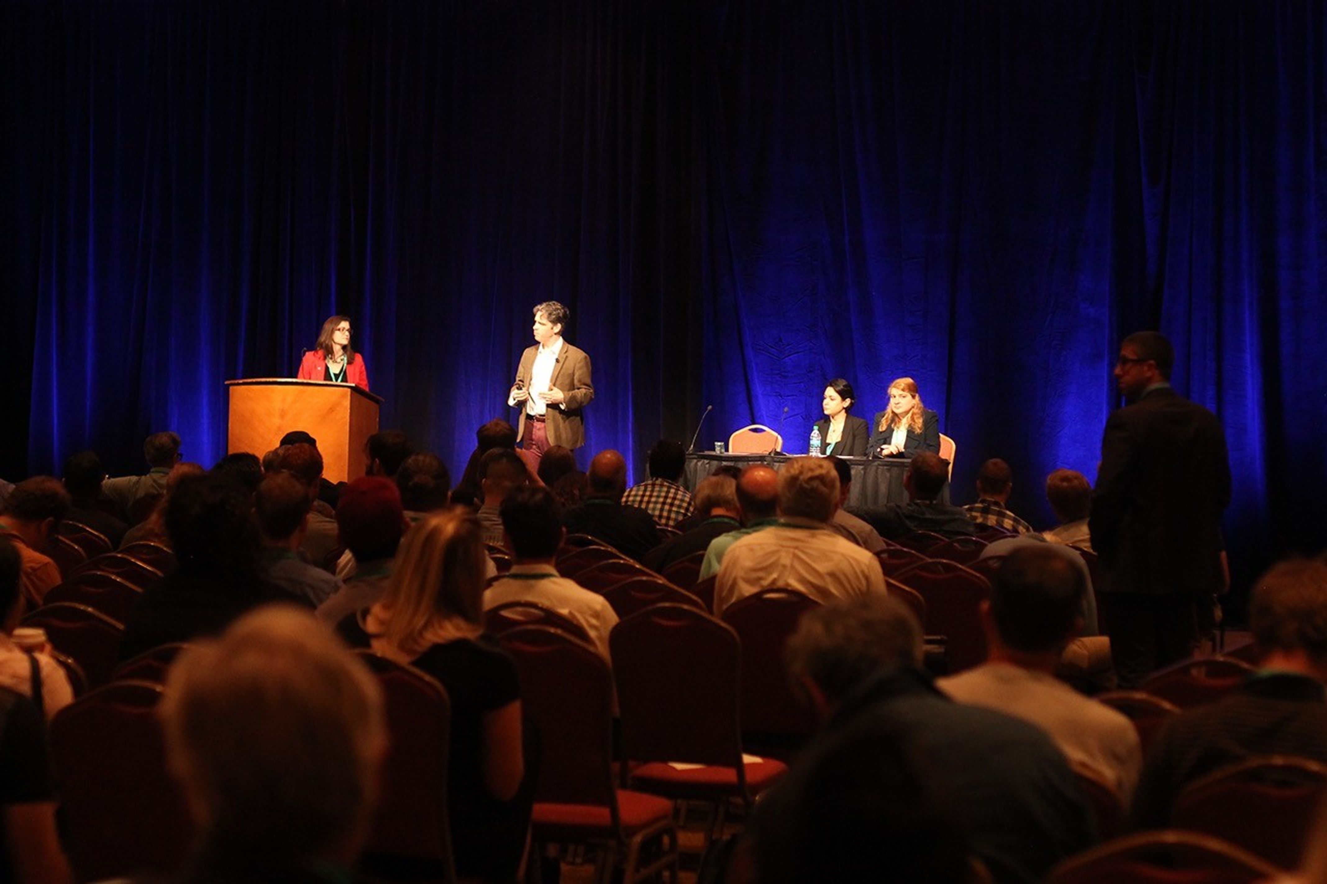 The backs of peoples' heads in the audience are seen in as they sit in their chairs watching the scientists on stage. One woman in a red jacket with dark hair stands behind a podium to the left. A man with dark hair and a brown suit jacket stands to her left speaking. Two women sit at a table to the right, one with dark hair and one with red hair, and both wearing black jackets.