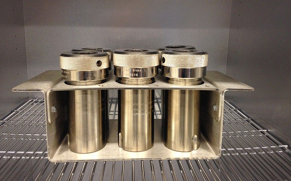 The image shows five (two rows with three in the front and two in the back with a gap between the two) golden metal cylinders containing samples on a wire shelf in a fridge. The canisters are capped with what appear to be screw-top metal lids. They are held is a rectangular casing of the same golden metal material.