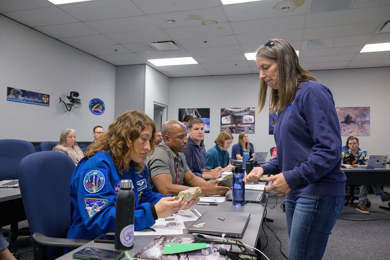 The Artemis crew sits at table to the right of frame. In the foreground, an astronaut with long curly hair dressed in a jumpsuit examines a rock. Juliane stands to the right dressed in jeans and a blue sweater with a NASA logo. Juliane is talking and appears to have her right hand closed around a smaller sample.