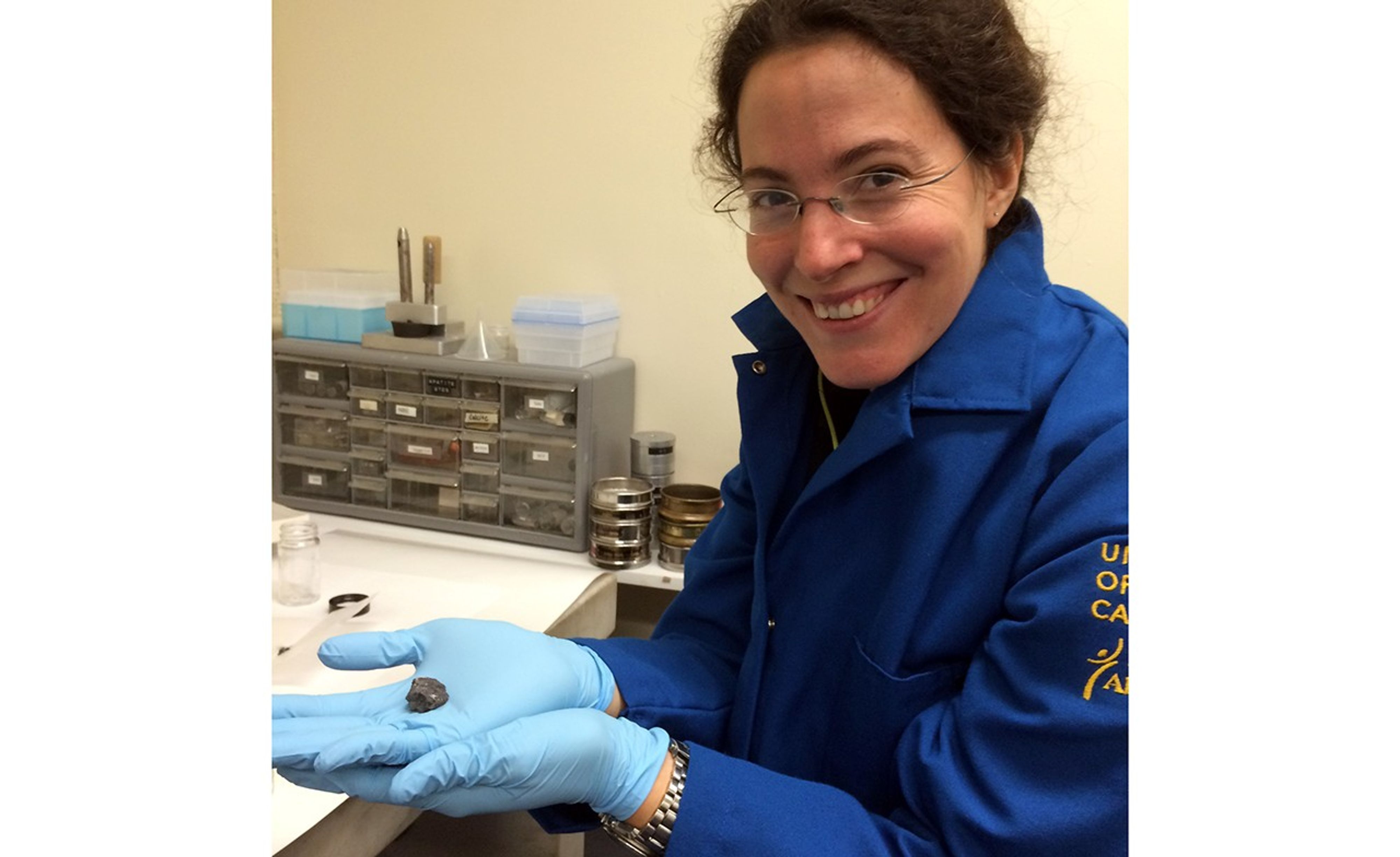 Melanie holds a small sample in two hands protected by blue, sterile, nitrile gloves. She smiles at the camera. Her hair is tied back and she wears glasses. She is sitting at a sample preparation desk.