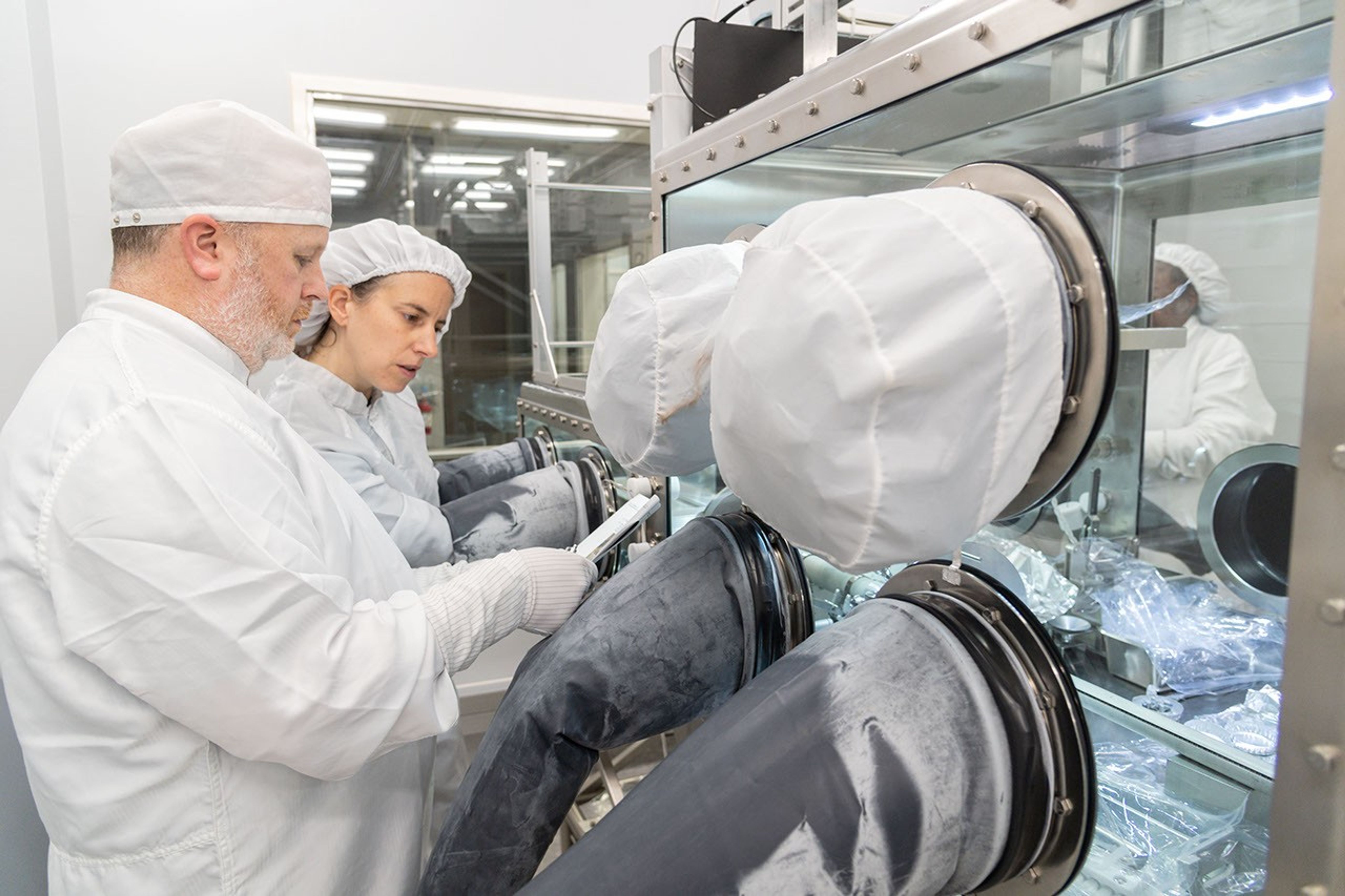 A woman in center frame has her hands inserted into the gloves of the sterile sample chamber. A man with a short beard to the left of frame holds out a paper in his hand so that she can read what is written as she works. Both are dressed in sterile laboratory garb with only their faces visible.