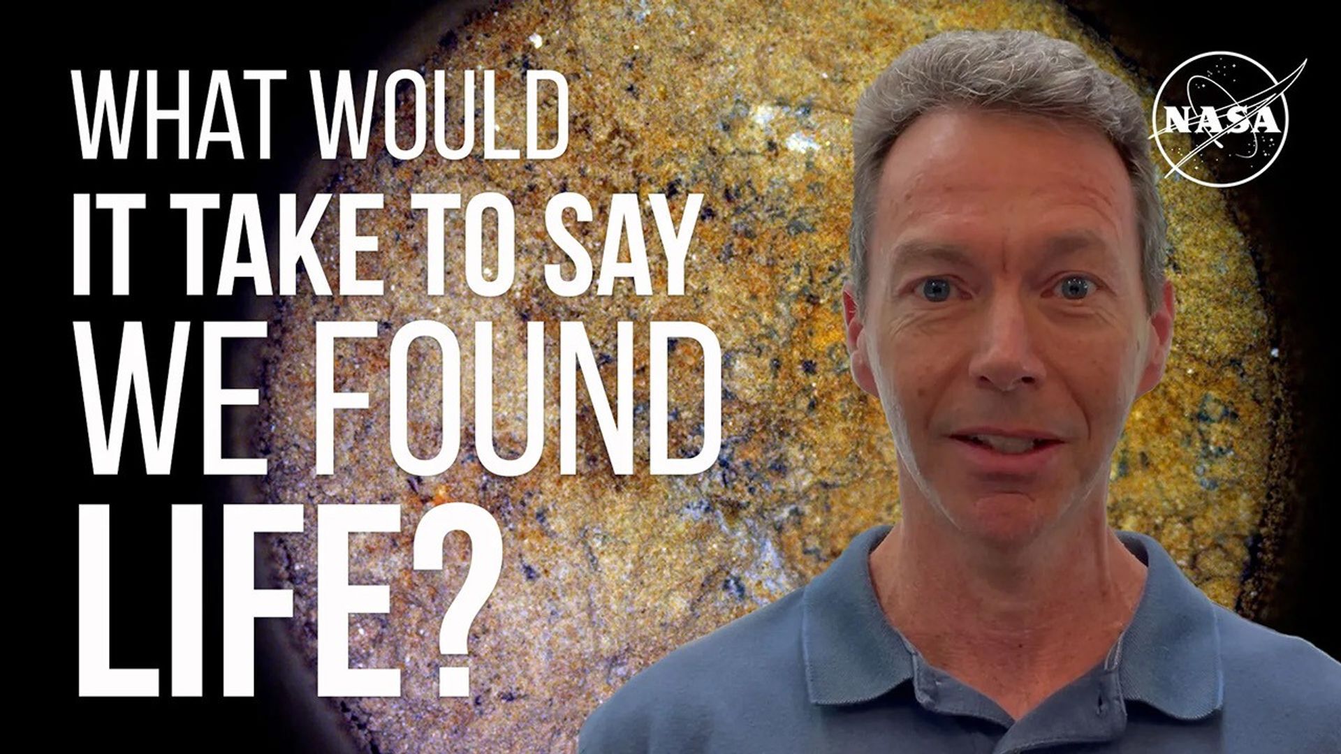 The image shows a man with blue eyes, grey/brown hair, and a blue shirt with collar looking at the camera. He is visible from shoulders up and stands to left of frame. To the right of frame are the words "What Would It Take to Say We Found Life?" To the top left is a white NASA meatball logo. In the background is a closeup image of a rock surface.