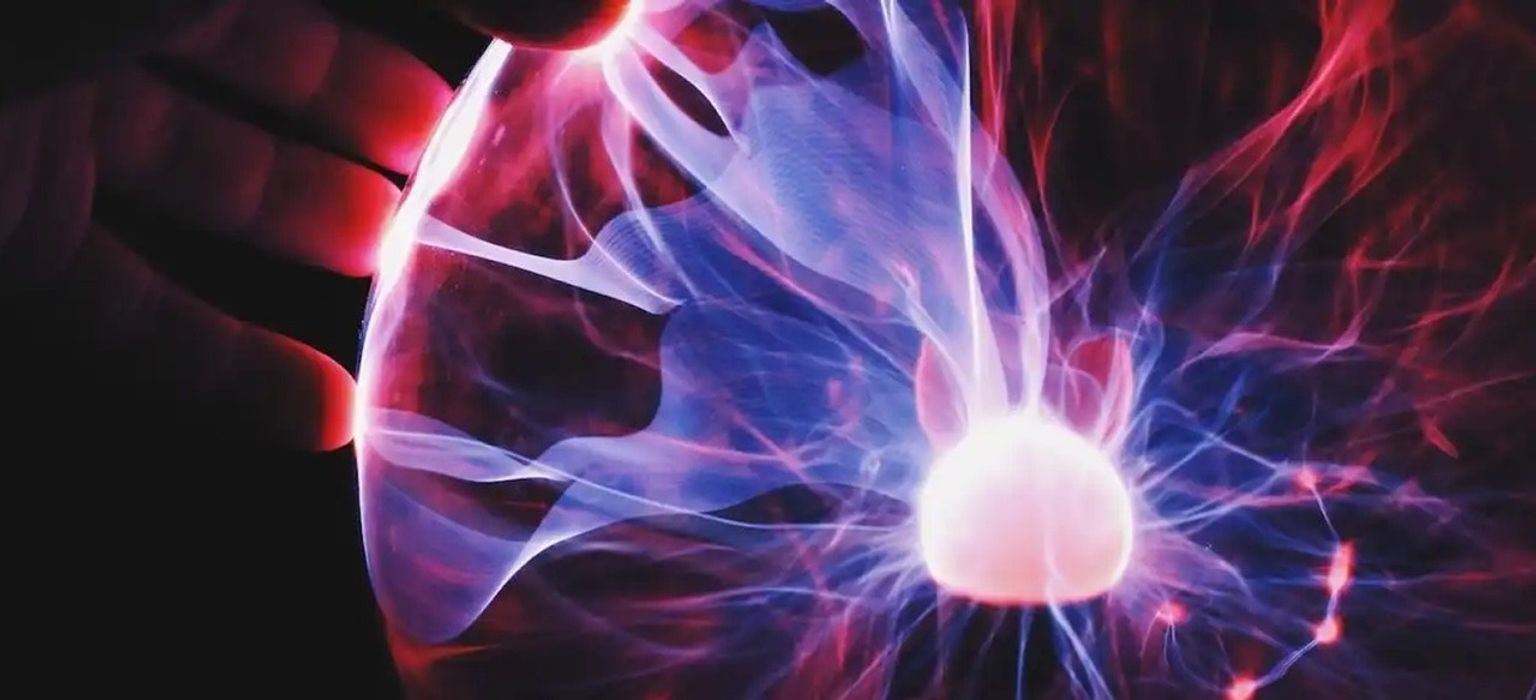 Close-up of a plasma ball. A hand is touching the surface of the glass sphere, and tendrils of pink and blue light are emanating from a central glowing orb and reaching outwards.