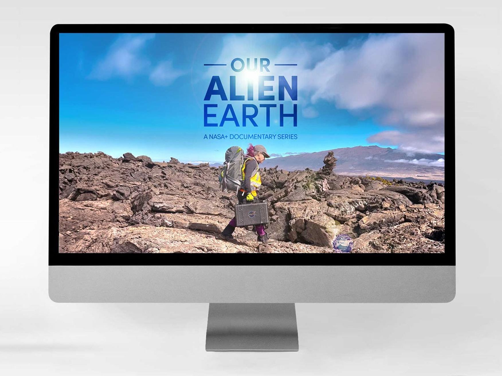 A desktop monitor displaying displaying a scene from Our Alien Earth featuring a NASA scientist dressed in field expedition clothing and carrying scientific equipment walking across lava fields in Hawai'i.