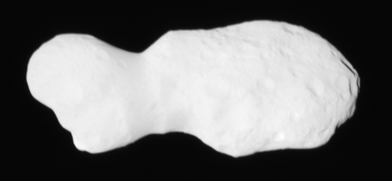 A light gray rocky object lies starkly against a pitch-black background. The object appears elongated with a narrower section on the left side and wider section on the right, resembling a peanut or dog bone shape. The surface appears rough and cratered with visible light and shadow areas highlighting its uneven texture.