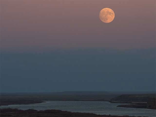 Photograph of a full Moon over a river and marshland. The background sky is turning dark, in hues of blue and pink.