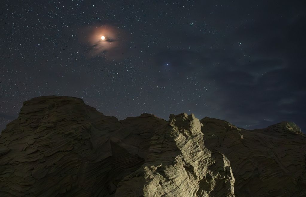 The eclipsed Moon glows red in a starry, partially hazy night sky, above a cliff of layered rock in the foreground.