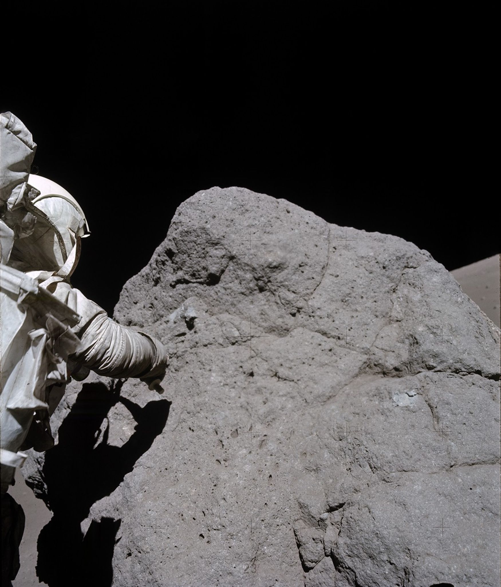 A person wearing a bulky, white spacesuit is partially visible on the left side of the image, extending a gloved hand toward a large, rough-textured, gray boulder. The boulder has a dusty, uneven surface with cracks and crevices. The person and boulder are lit up, while the background is mostly black. The person’s shadow provides a black shape cast on the boulder, highlighting the contours of both the boulder and the person.