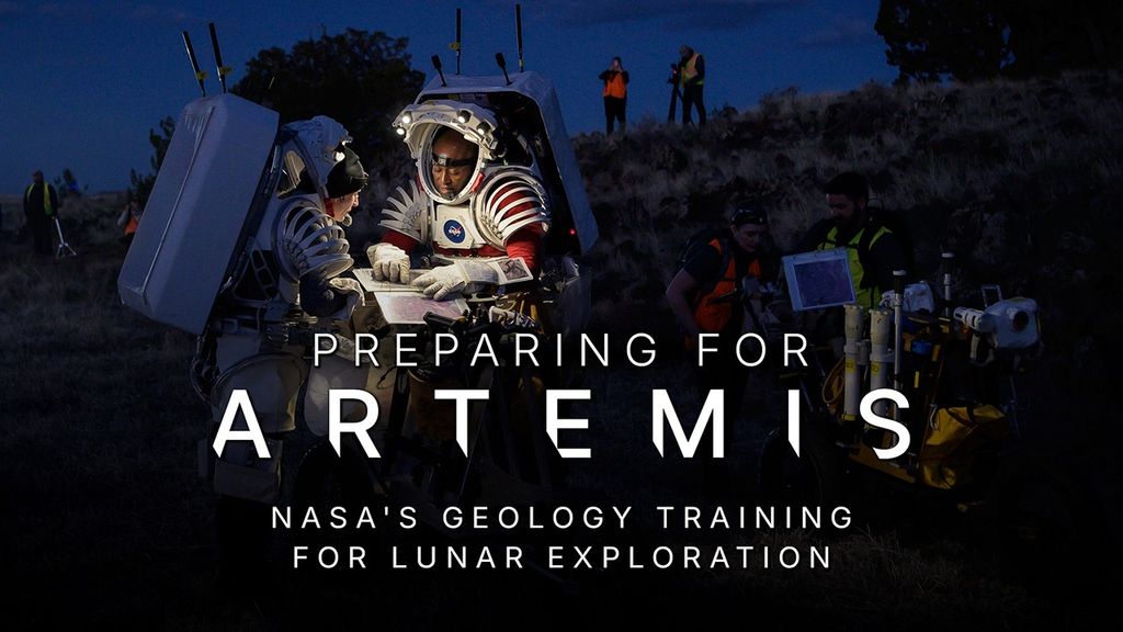 A nighttime picture of two astronauts wearing partial spacesuit mockups with large, rectangular backpacks. They are standing outside in a rocky terrain with trees in the background and are gathered around a cart with goldenrod, blue, and stainless steel-colored components on it. Small lights from the rims of their space suit “helmets” illuminate their work area. They are pointing to a map inside a three-ring binder. A few observers are standing in the background. Text reads: Preparing for Artemis - NASA's Geology Training for Lunar Exploration.