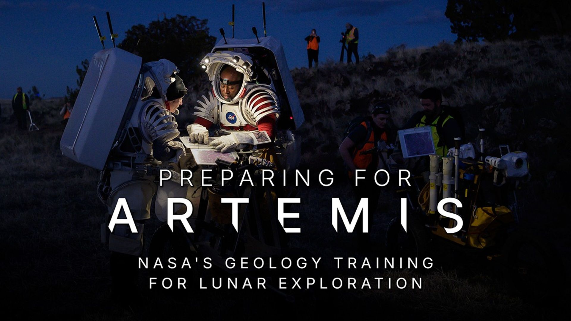 A nighttime picture of two astronauts wearing partial spacesuit mockups with large, rectangular backpacks. They are standing outside in a rocky terrain with trees in the background and are gathered around a cart with goldenrod, blue, and stainless steel-colored components on it. Small lights from the rims of their space suit “helmets” illuminate their work area. They are pointing to a map inside a three-ring binder. A few observers are standing in the background. Text reads: Preparing for Artemis - NASA's Geology Training for Lunar Exploration.
