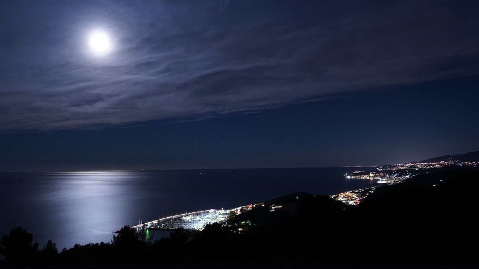 A bright Moon in the night sky reflects its light on the water of a coastal town, with lights illuminating the coast.