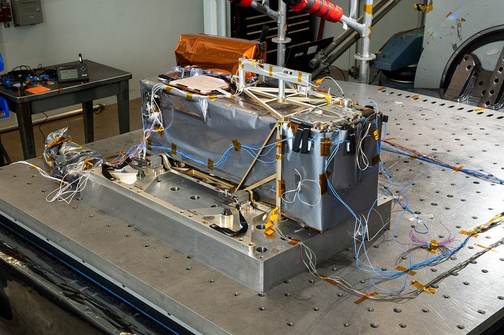Large metallic worktable with a rectangular machine-like object mounted on a metal base. The object is wrapped in silver and copper-colored material with exposed frames, brackets, and panels. Numerous thin blue, white, and multicolored wires extend from the sides and lie across the table surface. Additional equipment, metal supports, and tables appear in the background.