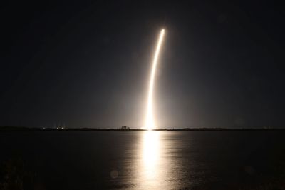 Fotografía de larga exposición del lanzamiento nocturno del cohete Firefly Aerospace, mostrando un brillante arco de luz mientras el cohete asciende hacia el cielo sobre una tranquila masa de agua con su resplandor reflejándose en la superficie.