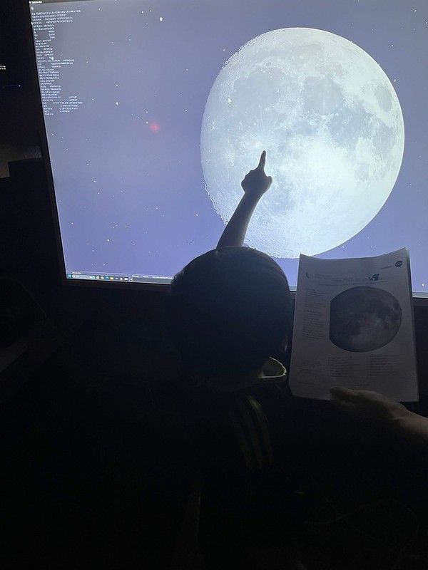 A young child points at a large waxing gibbous moon that's being shown on a large computer screen. A hand holds copies of Moon Maps next to the child.