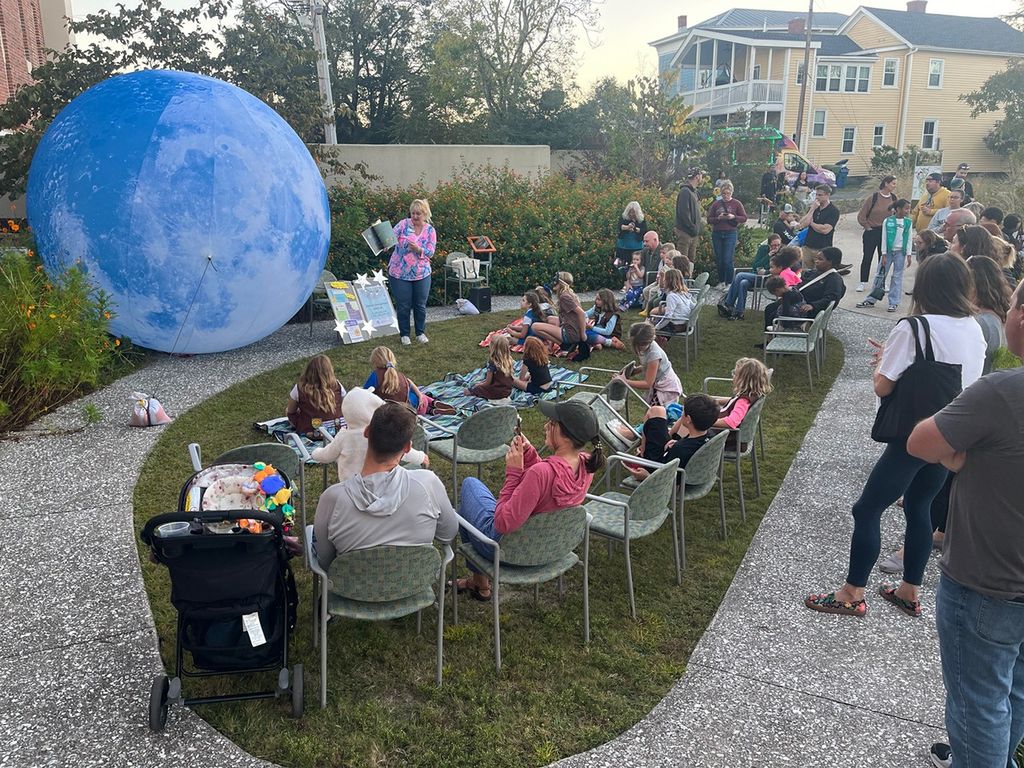 Librarian reads a story next to a gigantic Moon model, outdoors, with a crowd of families seated on chairs and on the grass and standing on the sidewalk nearby.