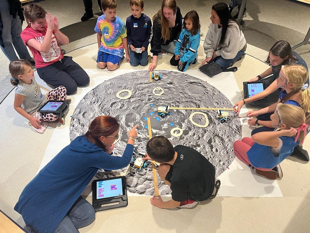 Crowd of families circled on the floor around an oversized map of the Moon, using tablets and pointing towards a model rover.