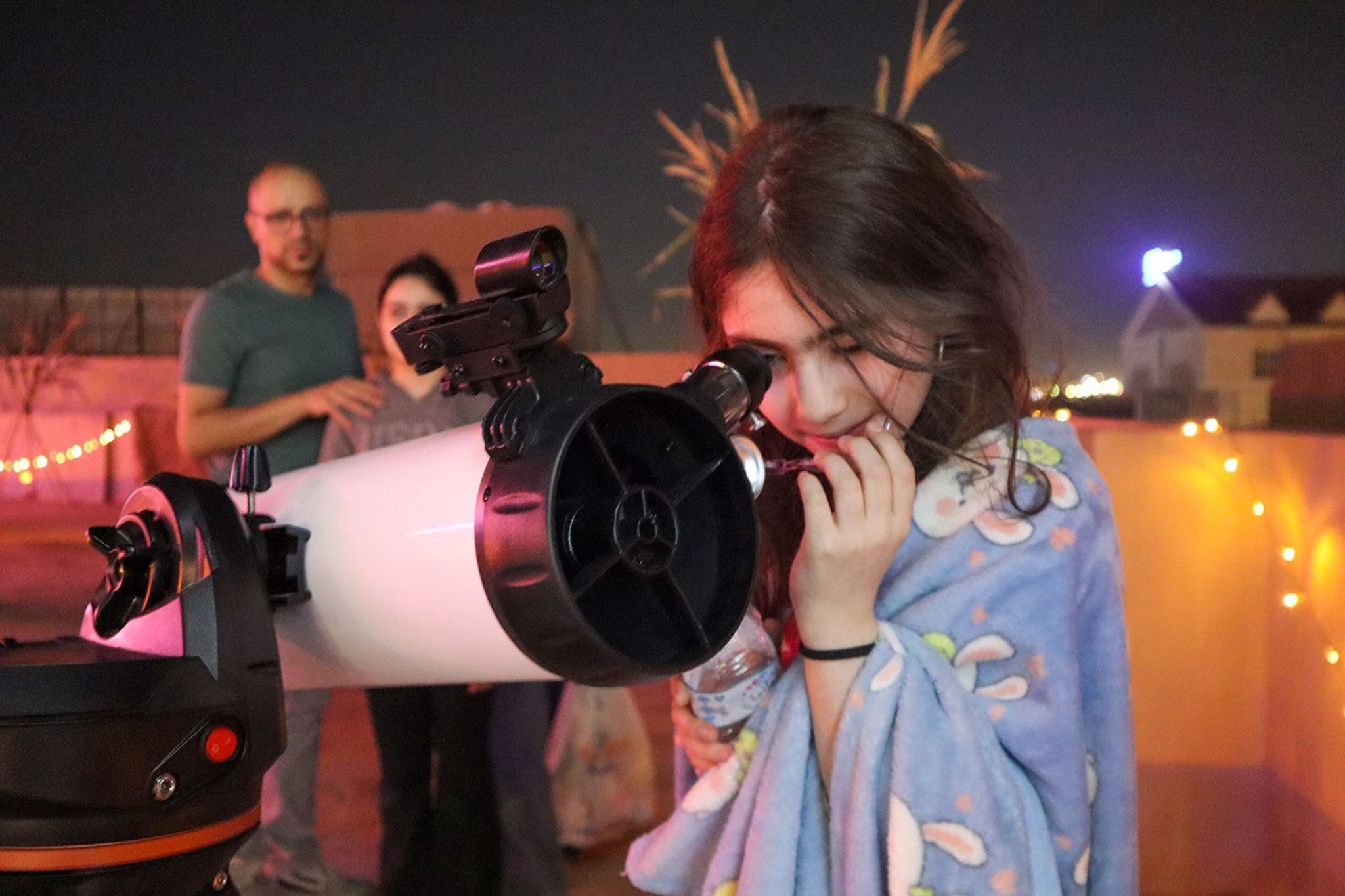 A young girl peers through a telescope while 2 people watch in the background.