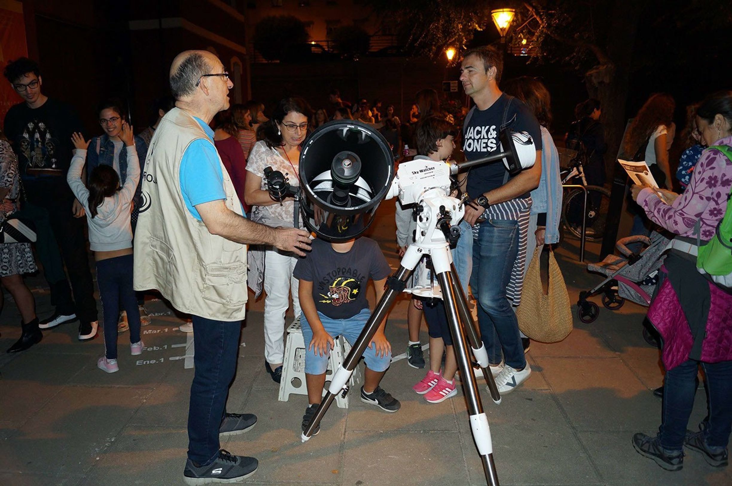 Two people have a conversation over a telescope, with other observers in the background.
